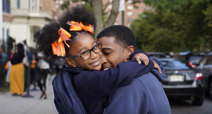 A father hugs his elementary school daughter, who is wearing glasses and orange bows, outside a Success Academy school.
