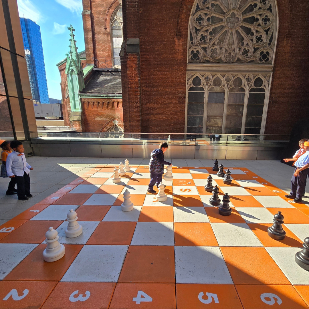 Our first graders are already thinking several moves ahead, playing chess on the rooftop with the city skyline behind them. See them at the board.